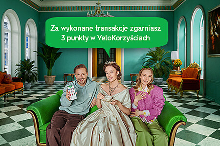 Mężczyzna, kobieta w sukience z epoki i młoda dziewczyna na zielonej sofie w pomieszczeniu z zielonymi roślinami i szachownicą na podłodze, reprezentują współczesność i przeszłość, nawiązując do spotu reklamowego kantoru walutowego.