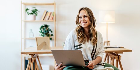 Młoda kobieta z laptopem siedzi uśmiechnięta w minimalistycznym pokoju.