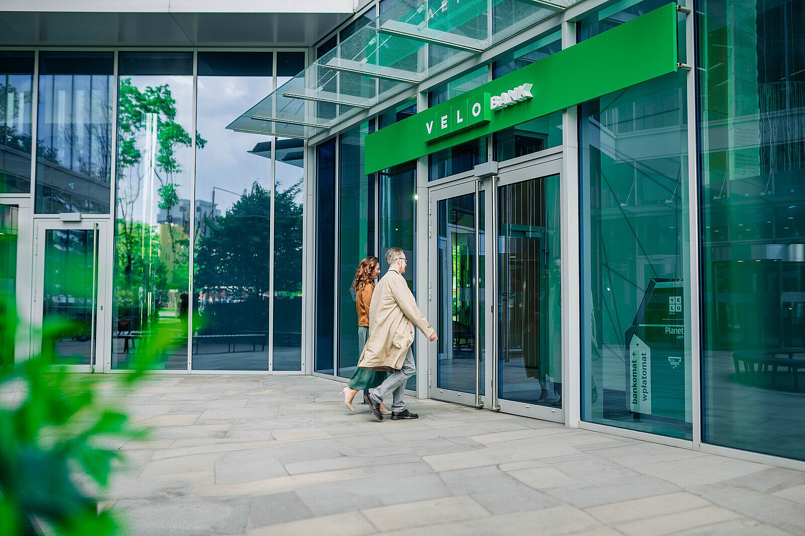 Dwoje ludzi w płaszczach, trzymających się za ręce, kieruje się do wejścia w zielonej elewacji banku Velo Bank w nowoczesnym budynku ze szklaną fasadą.