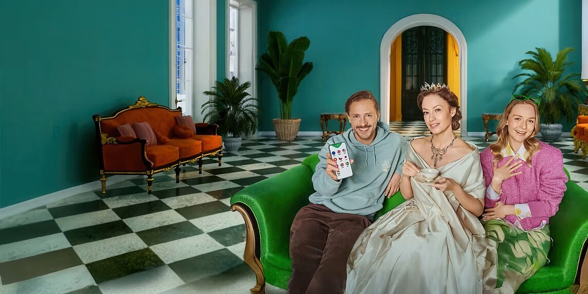 Mężczyzna, kobieta w sukience z epoki i młoda dziewczyna na zielonej sofie w pomieszczeniu z zielonymi roślinami i szachownicą na podłodze, reprezentują współczesność i przeszłość, nawiązując do spotu reklamowego kantoru walutowego.
