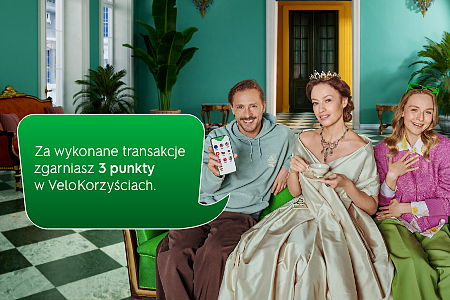 Mężczyzna, kobieta w sukience z epoki i młoda dziewczyna na zielonej sofie w pomieszczeniu z zielonymi roślinami i szachownicą na podłodze, reprezentują współczesność i przeszłość, nawiązując do spotu reklamowego kantoru walutowego.