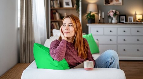 Kobieta pije kawę siedząc w swoim salonie i spokojnie patrzy przez okno