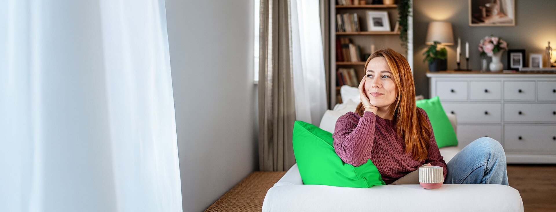 Kobieta pije kawę siedząc w swoim salonie i spokojnie patrzy przez okno