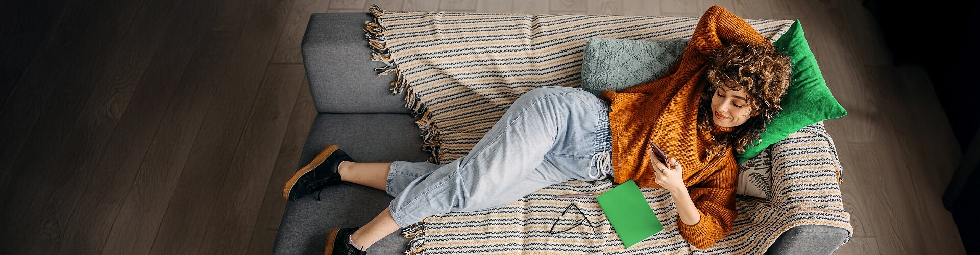 Dziewczyna w pomarańczowym swetrze leży wygodnie na kanapie i korzysta ze smartfona.